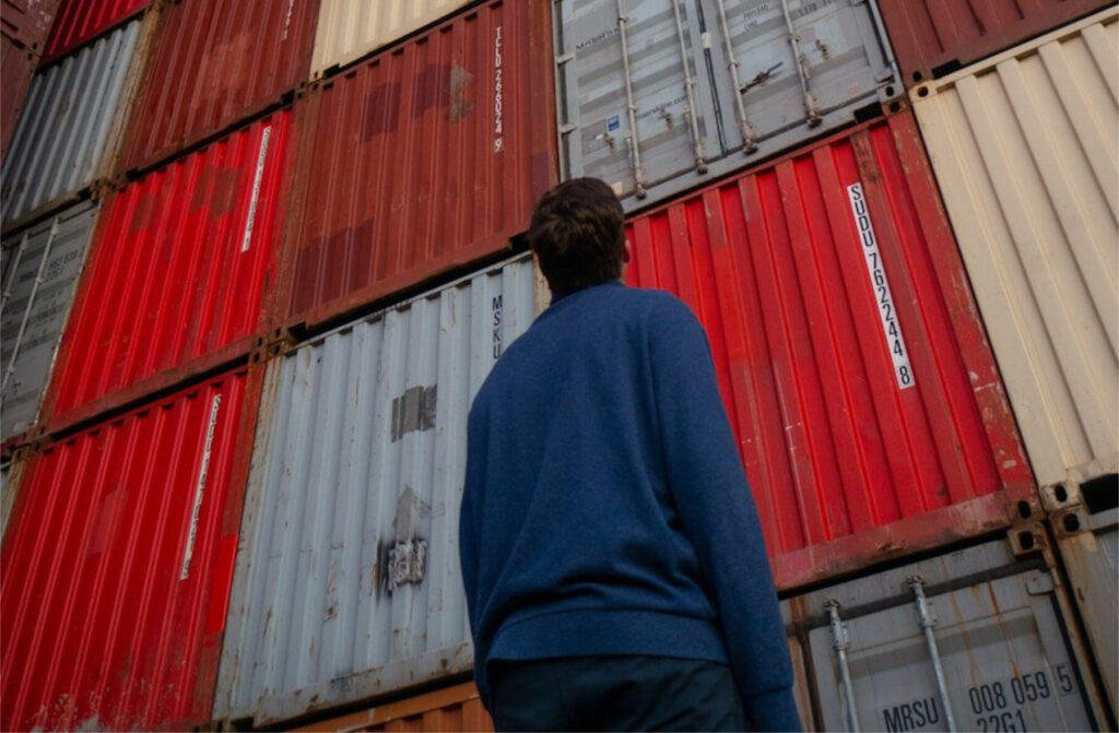 man in black jacket standing in front of red and blue intermodal containers
